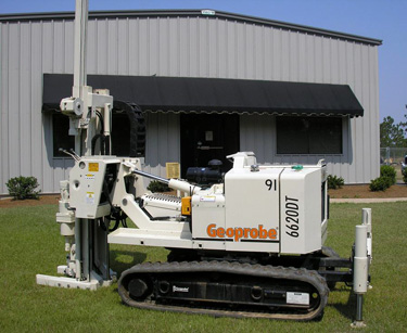 White and orange Geoprobe 6620DT drilling machine on lawn in front of industrial building