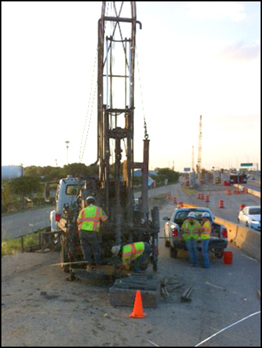 Construction crew operating drilling rig on paved road with safety equipment