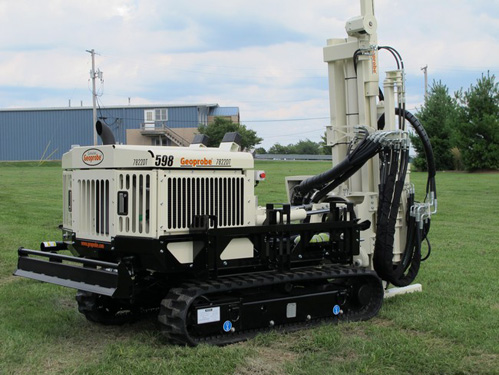 White track-mounted Geoprobe drilling rig on grassy field with drilling mast extended