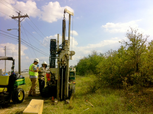 Two utility workers in safety vests operating drilling rig on rural roadside